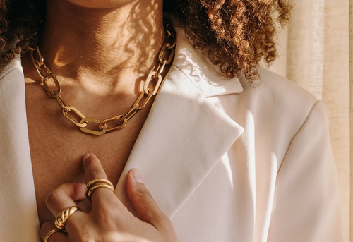 A person wearing a white blazer, a chunky gold chain necklace, and gold rings touches their chest.