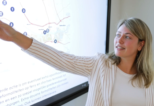A woman in a striped blazer points to a map on a screen that shows routes and locations like Rotterdam.