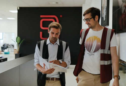 Two men in reflective vests walk and review a document.