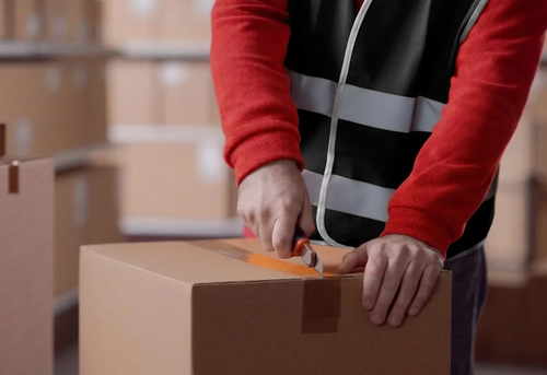 A person in a reflective vest uses a box cutter to open a sealed cardboard box in a warehouse with stacked boxes in the background.