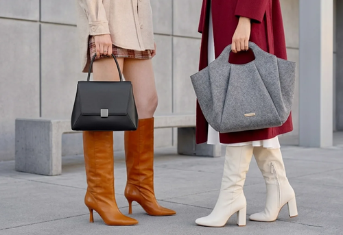 Two women stand on a sidewalk, one wearing tan boots and holding a black handbag, the other in white boots holding a large gray bag.