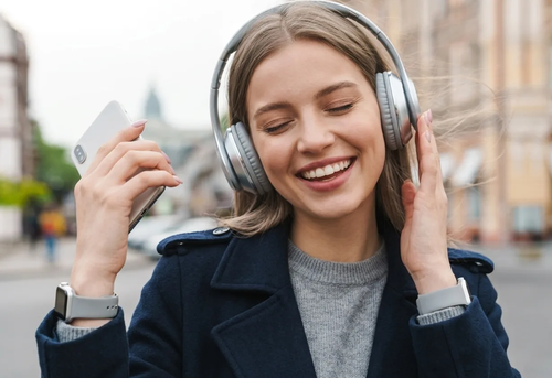 A young woman wearing headphones smiles with her eyes closed while holding a smartphone outdoors.