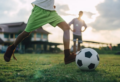 A child in green shorts prepares to kick a soccer ball on a grassy field, with an adult standing in the background.