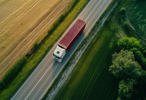 A white semi-truck with a red trailer drives on a rural road bordered by fields and trees, viewed from above.