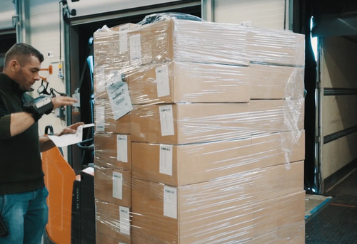 A man uses a scanner to check a stack of shrink-wrapped boxes next to a forklift.