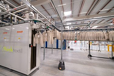 Jackets on hangers move along an automated conveyor system in an industrial setting with metal pipes and equipment.
