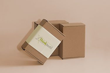 Three stacked brown cardboard boxes with a "Thinktextil" label on the frontmost box against a beige background.