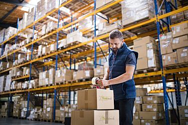 A man is sealing cardboard boxes with tape in a large warehouse with shelves filled with various boxed goods.