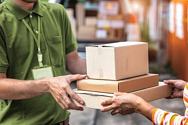 A delivery person in a green shirt hands over three stacked cardboard boxes to another person.