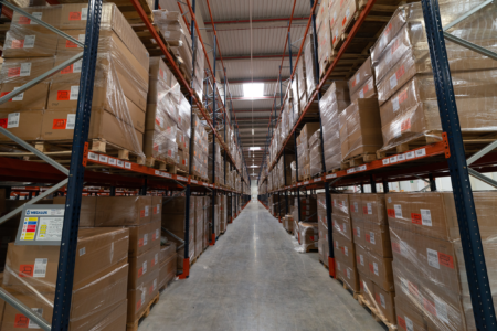 Interior of a warehouse with tall shelves stocked with pallets of boxes, extending into the distance under fluorescent lighting.