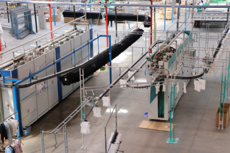 Industrial laundry facility with automated clothing conveyor system. Hanging white and dark garments move along overhead tracks in a large, spacious environment.