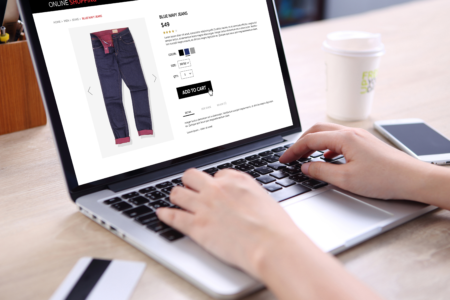 Person browsing an online shopping site on a laptop, looking at blue mint jeans priced at $49, with white and navy color options. A coffee cup is on the right.