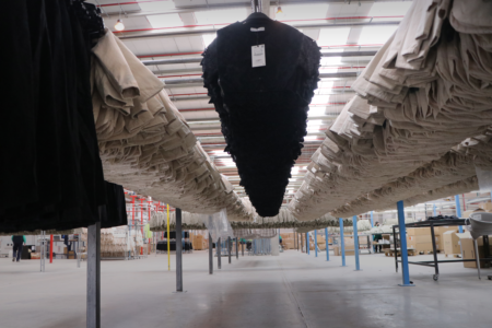 Interior of a clothing factory with garments hanging on overhead conveyors. Black and beige items are organized in rows, stretching into the distance under bright overhead lights.