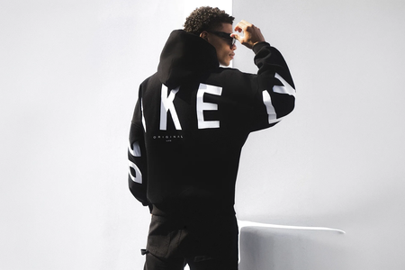 A person wearing a black hoodie with large white letters and black pants stands facing away, adjusting sunglasses in a minimal, well-lit room.