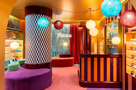 A brightly colored boutique interior with checkerboard and striped patterns, round hanging lamps, velvet furniture, and a neon "Fabulous" sign on the wall.