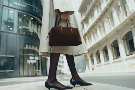 Person in a light-colored trench coat and black heels holding a dark brown handbag while standing on a city street.