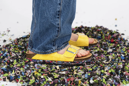A person wearing yellow sandals and blue jeans stands on a pile of colorful shredded material scattered on a white surface.