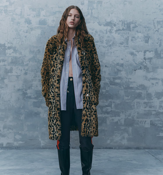 Person wearing a leopard print coat over a striped shirt, black pants, and black knee-high boots stands against a gray textured background.