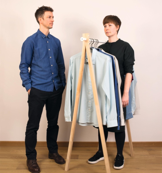 Two people stand beside a wooden clothing rack with shirts. The person on the left wears a blue shirt and black pants. The person on the right wears a black top and looks at the other person.