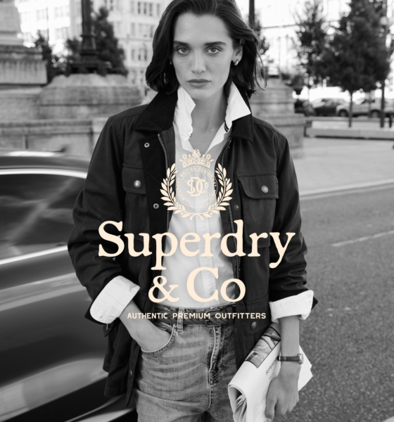 A woman stands on a city street holding a newspaper, wearing a jacket and collared shirt. The Superdry & Co logo and "Authentic Premium Outfitters" text are overlaid on the image.