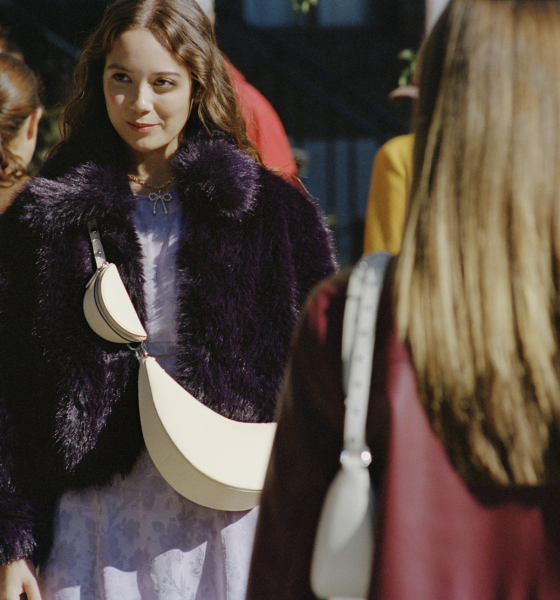 A woman in a dark fur coat and holding a white bag stands outdoors, facing another woman with long hair and a white bag. Several people are in the background.