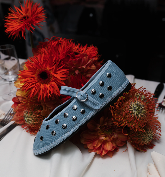 A light blue Mary Jane shoe with metallic studs rests on orange flowers, arranged on a white tablecloth with silverware and glassware nearby.