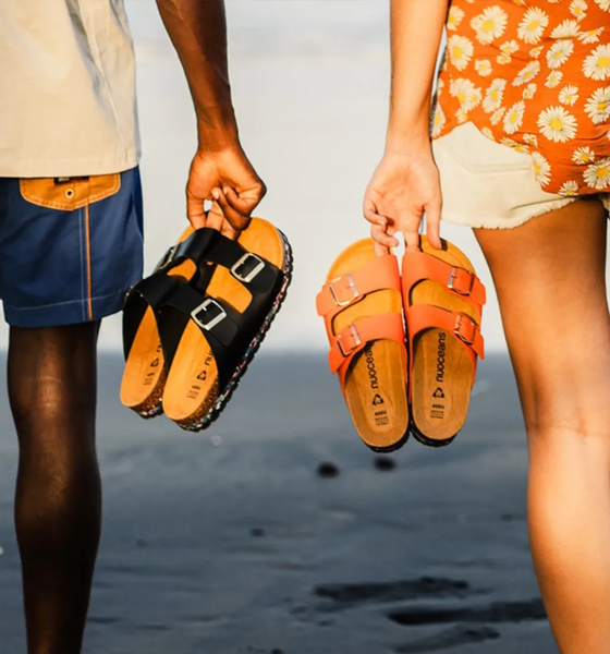 Two people stand side by side on a beach, holding sandals in their hands; one person wears blue shorts, the other wears white shorts and a floral top.