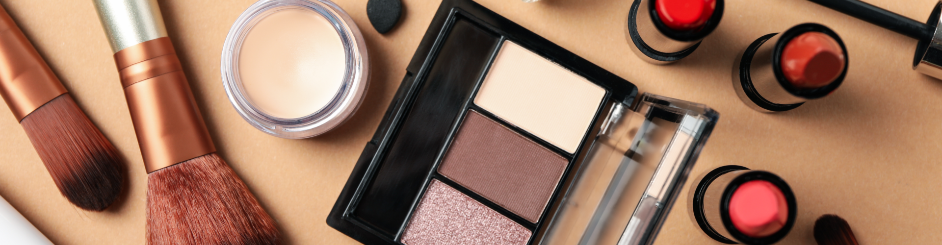 A variety of makeup products including brushes, lipsticks, nail polish, eyeshadow palette, foundation, and concealer arranged on a beige background.