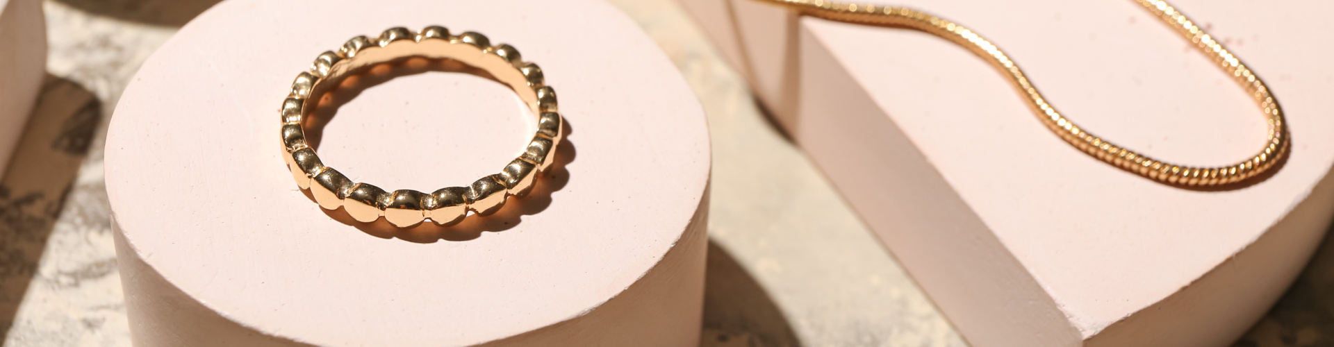 Gold beaded bracelet and gold chain necklace displayed on white geometric platforms.