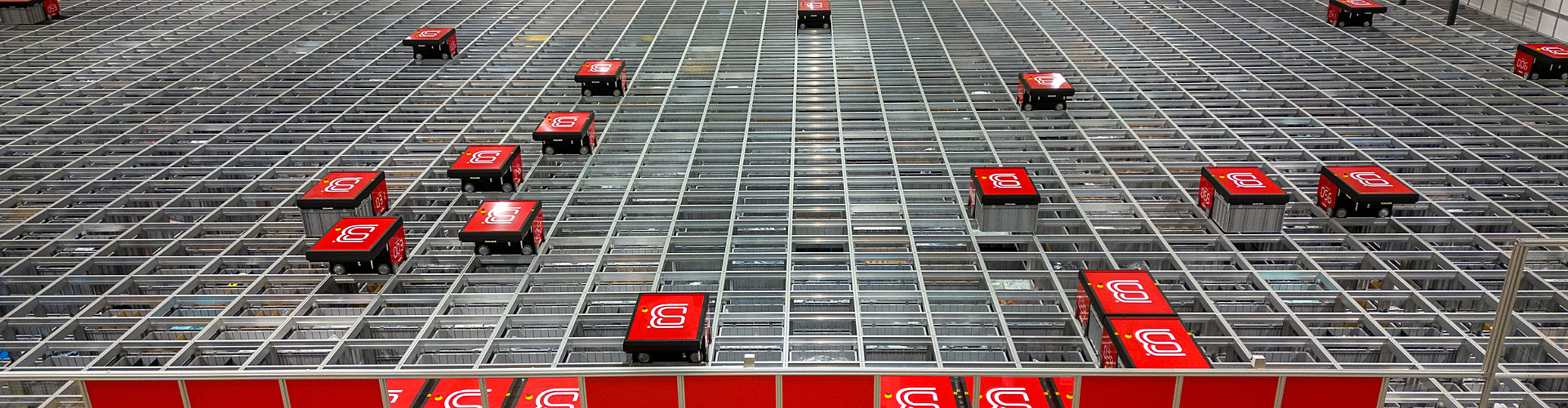 Automated robots with red covers move along a grid system in a large warehouse, likely sorting or retrieving goods for inventory management or order fulfillment.