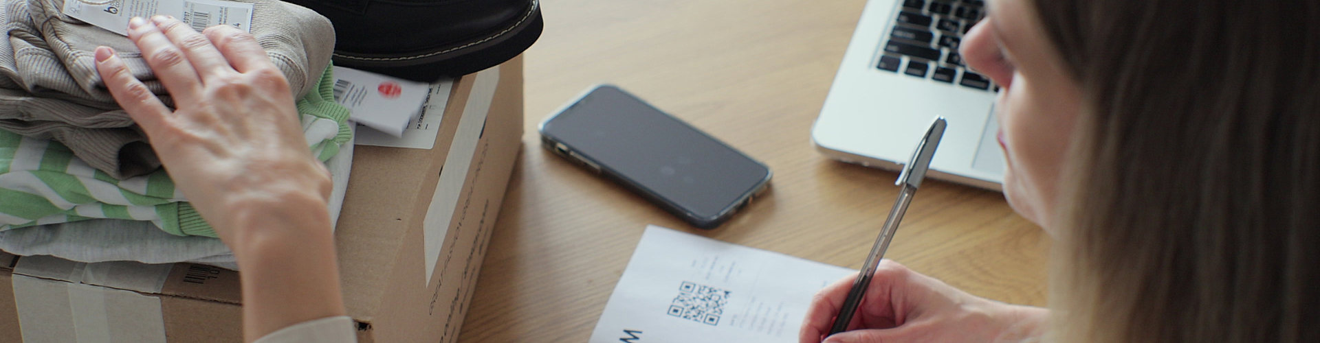 A woman fills out a return form next to a box with clothes and shoes, a smartphone, and a laptop on a wooden table.