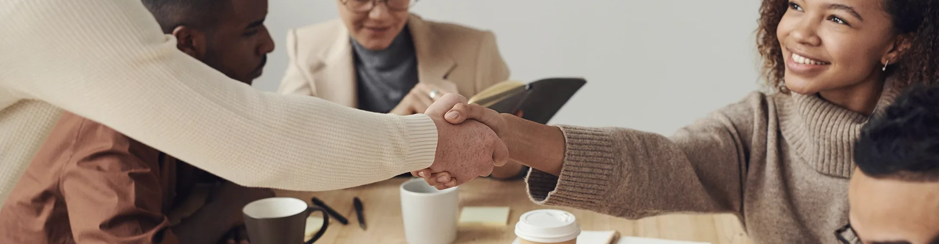 Two people shake hands across a desk while others work and take notes nearby in an office meeting setting with coffee cups and notebooks on the table.