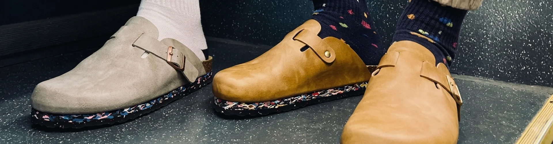 Two people wearing different colored clogs with patterned soles; one wears white socks and grey shoes, the other wears dark socks with tan shoes.