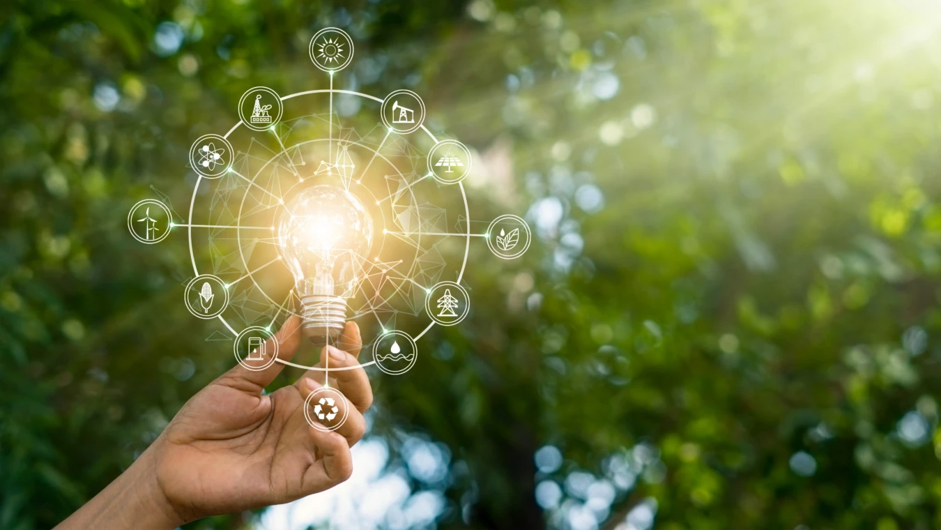 Hand holding a lit lightbulb surrounded by icons representing sustainability and technology against a green, blurred natural background.