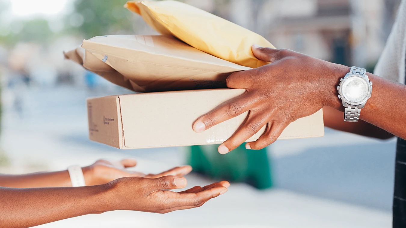 Two individuals exchanging parcels, with one person handing over a box and envelopes to another, against a blurred outdoor background.
