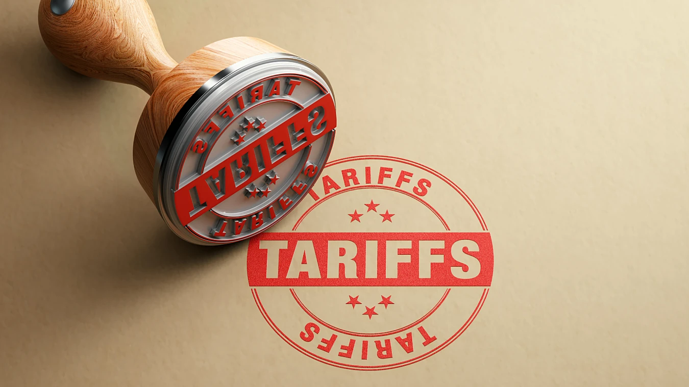A wooden stamp labeled "TARIFFS" lies next to a red-ink stamp imprint of the same word and design on a beige surface.