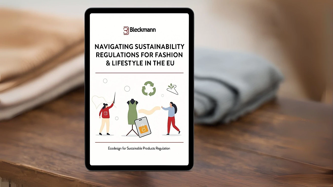 A tablet displays a document titled "Navigating Sustainability Regulations for Fashion & Lifestyle in the EU" by Bleckmann, resting on a wooden surface with folded clothes in the background.