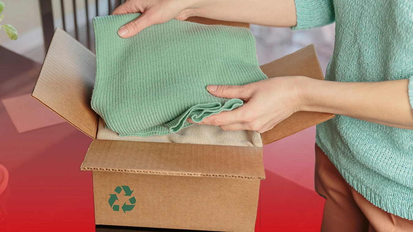 A person places a folded green sweater into a cardboard box with a recycling symbol printed on it.