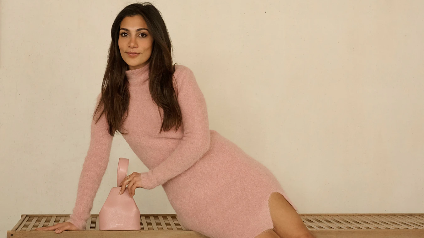 A woman in a pink long-sleeve sweater dress sits on a bench, holding a small pink handbag, against a plain beige background.