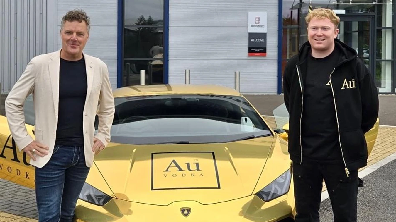 Two men stand in front of a gold Lamborghini with "Au Vodka" branding on the hood, parked outside a modern building.