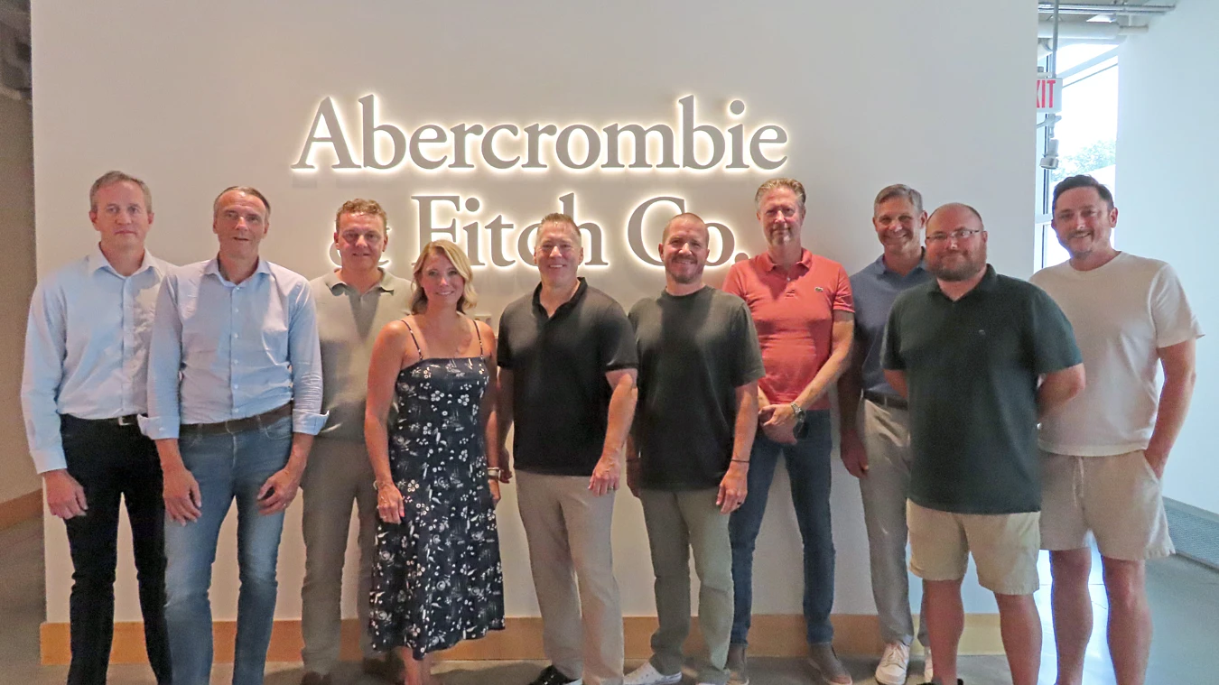 Ten people stand in front of an illuminated “Abercrombie Fitch Co.” sign inside an office space, facing the camera.