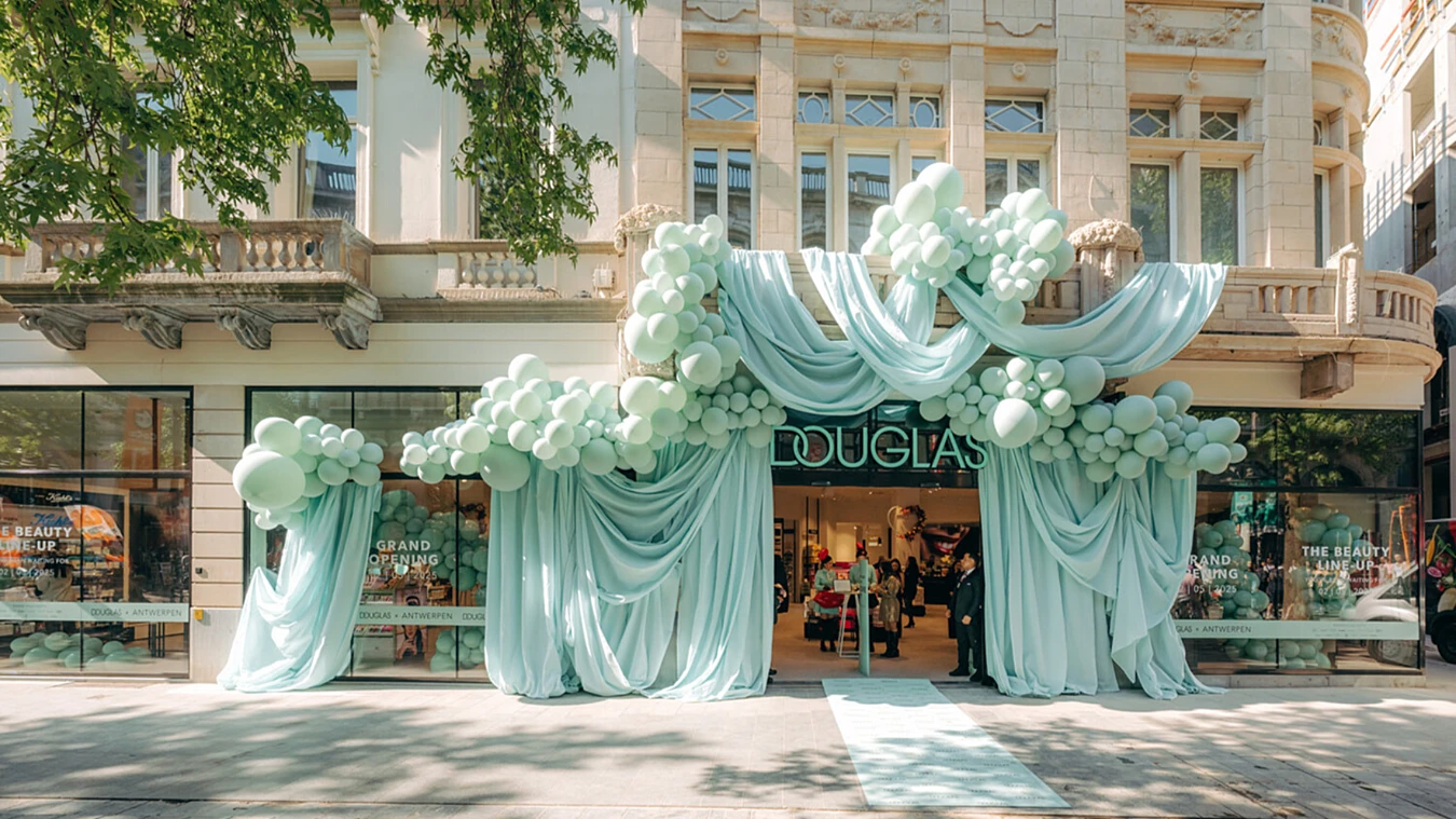 Storefront decorated with large clusters of light blue balloons and draped fabric, with “Douglas” sign above the entrance and people visible inside the store.