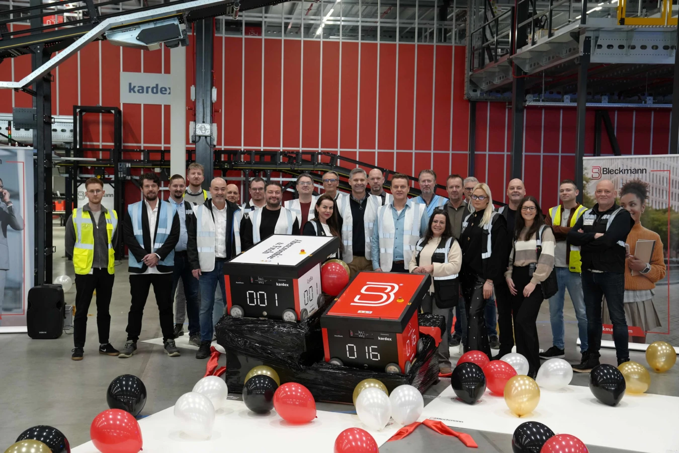 A group of people stands in a warehouse, posing behind two large boxes labeled “001” and “016,” surrounded by red, black, white, and gold balloons.