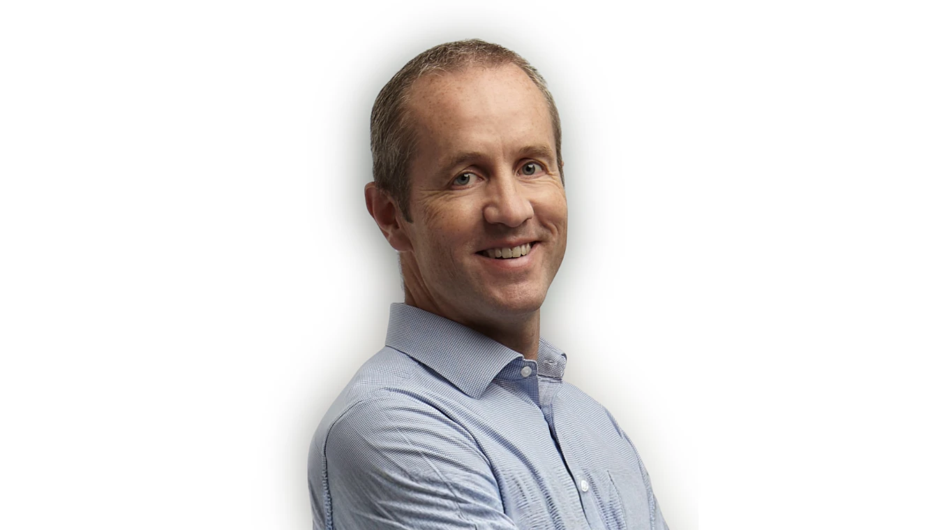 A middle-aged man with short hair wearing a light blue collared shirt stands with arms crossed and smiles at the camera against a white background.