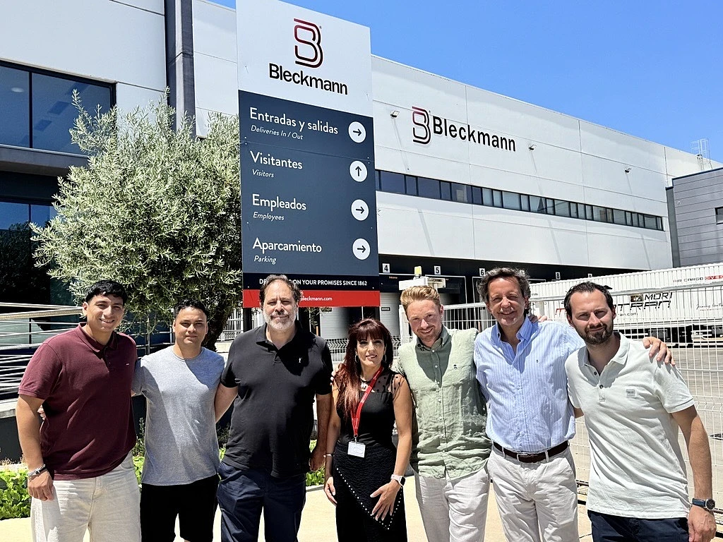 Six people stand in front of a Bleckmann building and sign on a sunny day, posing for a group photo outdoors.