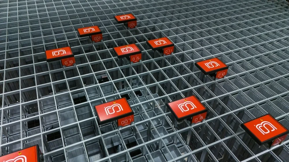 Red automated robots move along a metal grid system in a warehouse, likely part of an automated storage and retrieval system.