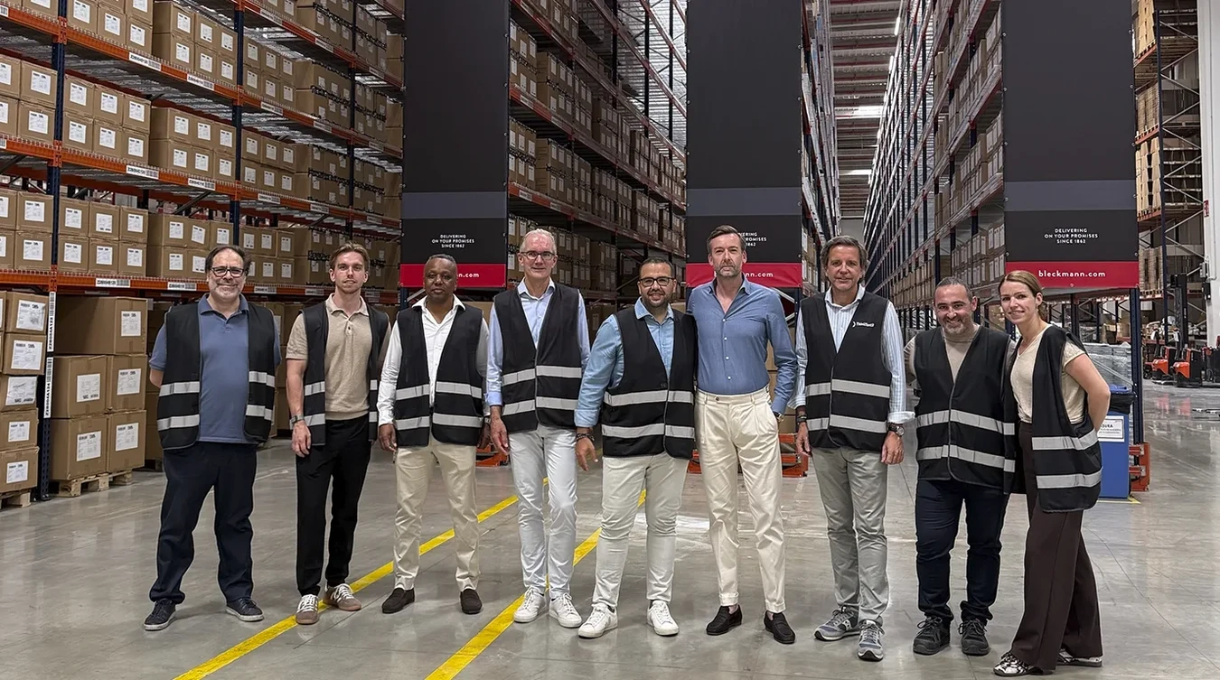 Nine people wearing black safety vests stand in a row inside a large warehouse with tall shelves filled with boxes.