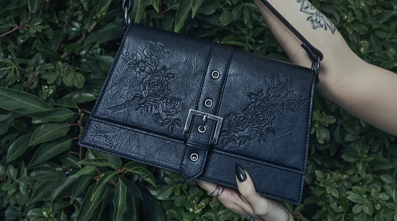 A person with black nail polish holds a black leather handbag with floral embossing and a front buckle, against a backdrop of green leaves.