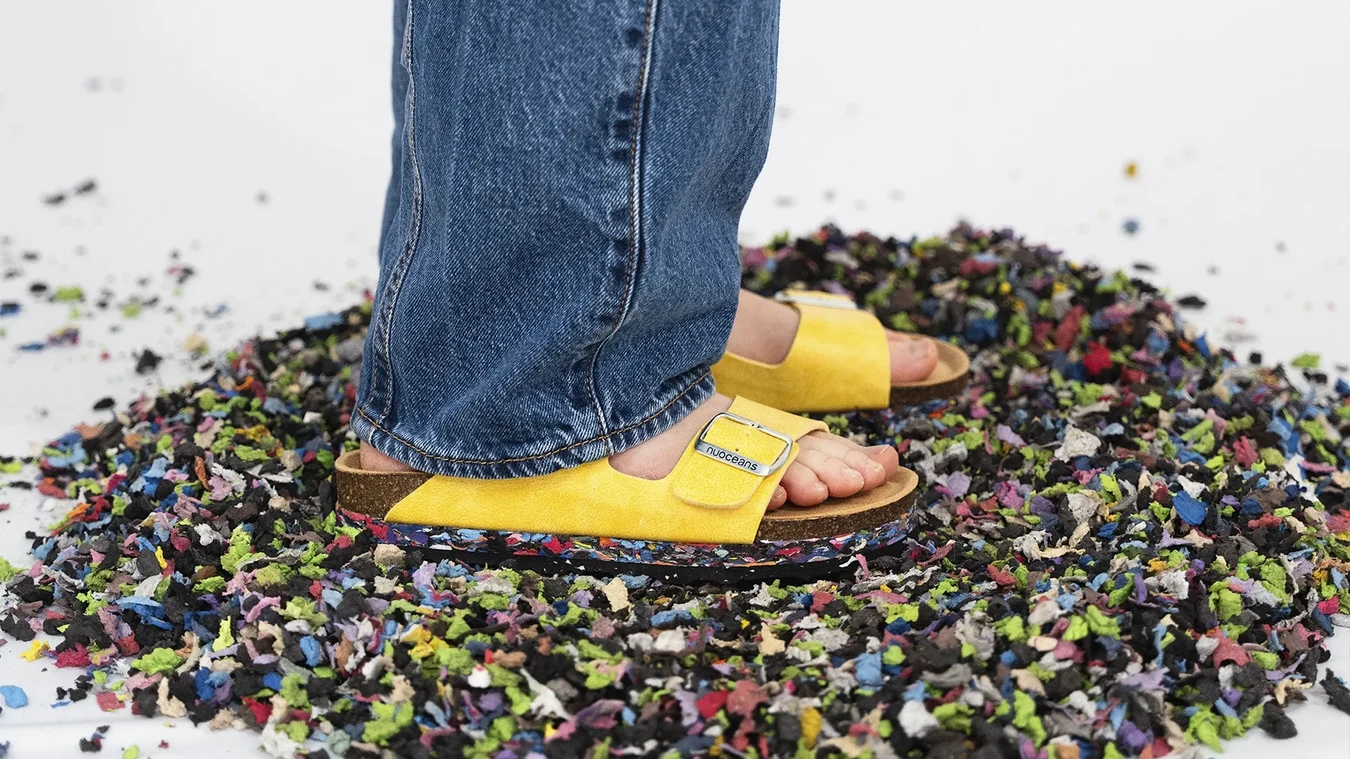 A person wearing yellow sandals and blue jeans stands on a pile of colorful shredded material scattered on a white surface.