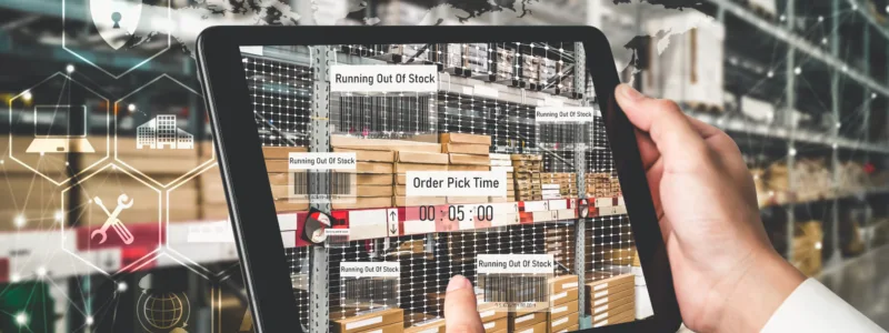 A person holds a tablet displaying inventory data in a warehouse, showing items running out of stock and order pick times, with shelves of boxes in the background.